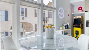Ein modernes Zimmer mit Glastisch, weißen Stühlen, einer Vase mit Blumen und einem Pac-Man-Spielautomaten, mit Blick auf die Straße.