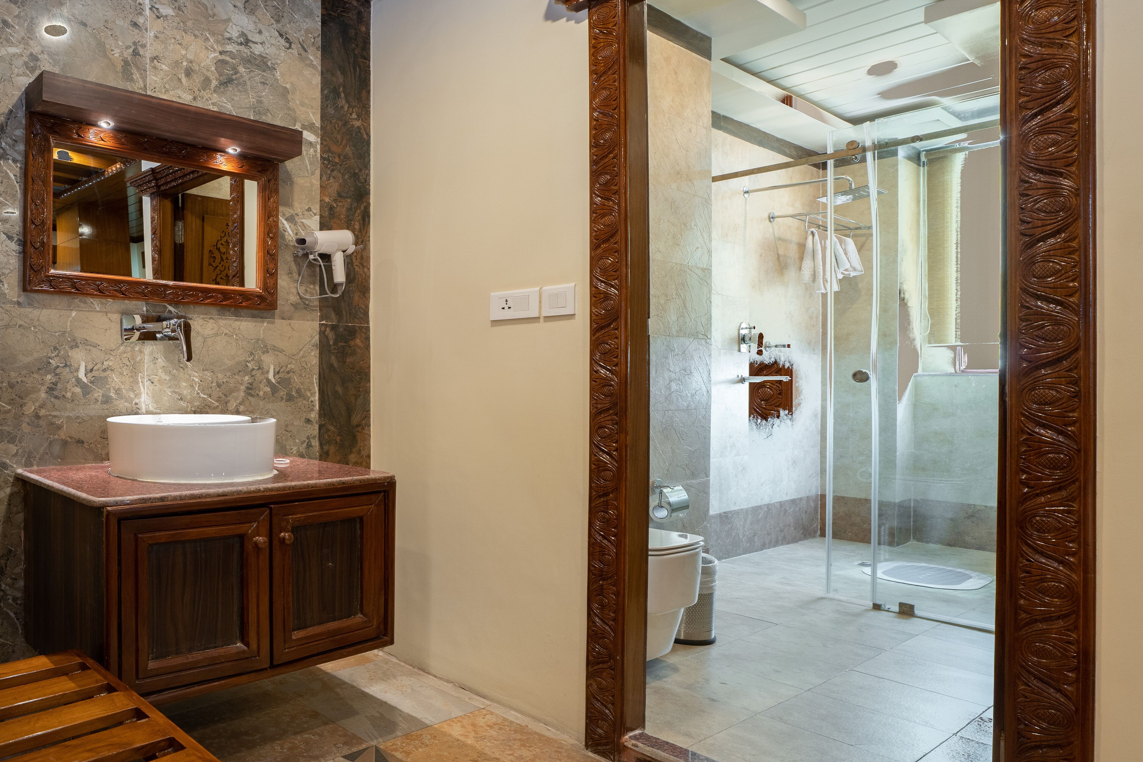 Une salle de bain avec un lavabo en marbre, des meubles en bois, un grand miroir et une douche séparée par une paroi de verre.