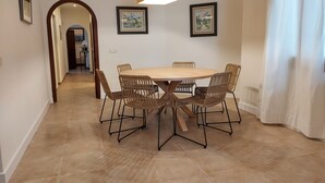 A dining area with a round table and six wicker chairs, a wooden floor, and two framed pictures on the wall.