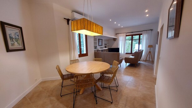 A dining area with a round table and six chairs, a hanging light fixture, and a kitchen in the background.