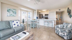 A living room with a sofa, a coffee table, and a patterned armchair. There is a dining area with a table and chairs. The kitchen is visible with a refrigerator, microwave, and cabinets. - Coral Penthouse | Corner Unit | Ocean Pointe 4216 (Tavernier)