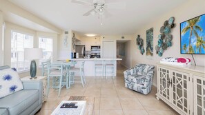 A living room with a sofa, a dining area with chairs, a kitchen with cabinets, a ceiling fan, and a TV on the wall. - Coral Penthouse | Corner Unit | Ocean Pointe 4216 (Tavernier)