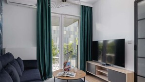 A modern living room with a dark sofa, a round coffee table with a magazine, a flat-screen TV on a wooden stand, and a balcony with green curtains.