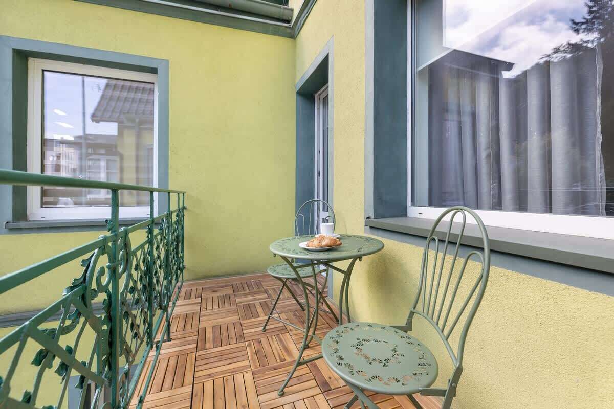 A balcony with a wooden floor, a metal railing, a small table with a chair, and a window with a view of a building and trees.