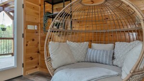 A wicker hanging chair with cushions in a wooden cabin with a window and a shelf with plants. - Southern Smoke | Scenic Decks | Hot Tub | Wi-Fi (Rogers)