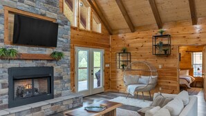 A cozy log cabin interior with a stone fireplace, a large TV mounted on the wall, and a comfortable seating area with a sofa and a hanging chair. - Southern Smoke | Scenic Decks | Hot Tub | Wi-Fi (Rogers)