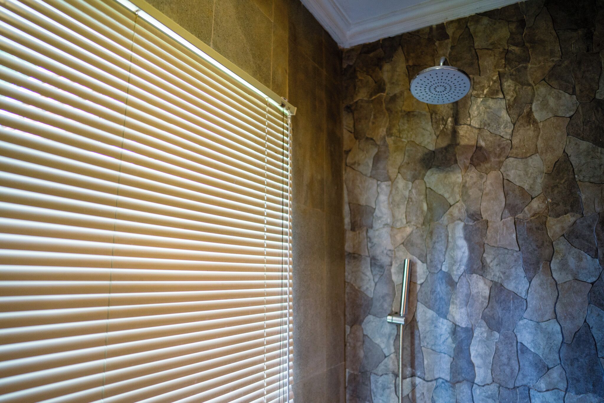 A bathroom with stone walls, a shower with a circular drain, and a window with horizontal blinds.