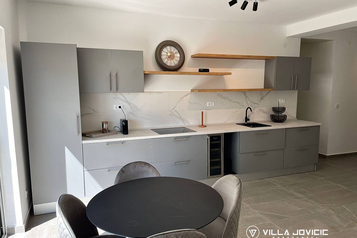 Een moderne keuken met grijze kastjes, een marmeren aanrechtblad en een ronde eettafel met stoelen.