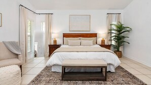 A bedroom with a large bed, bedside tables, a bench, a potted plant, and a framed picture on the wall.