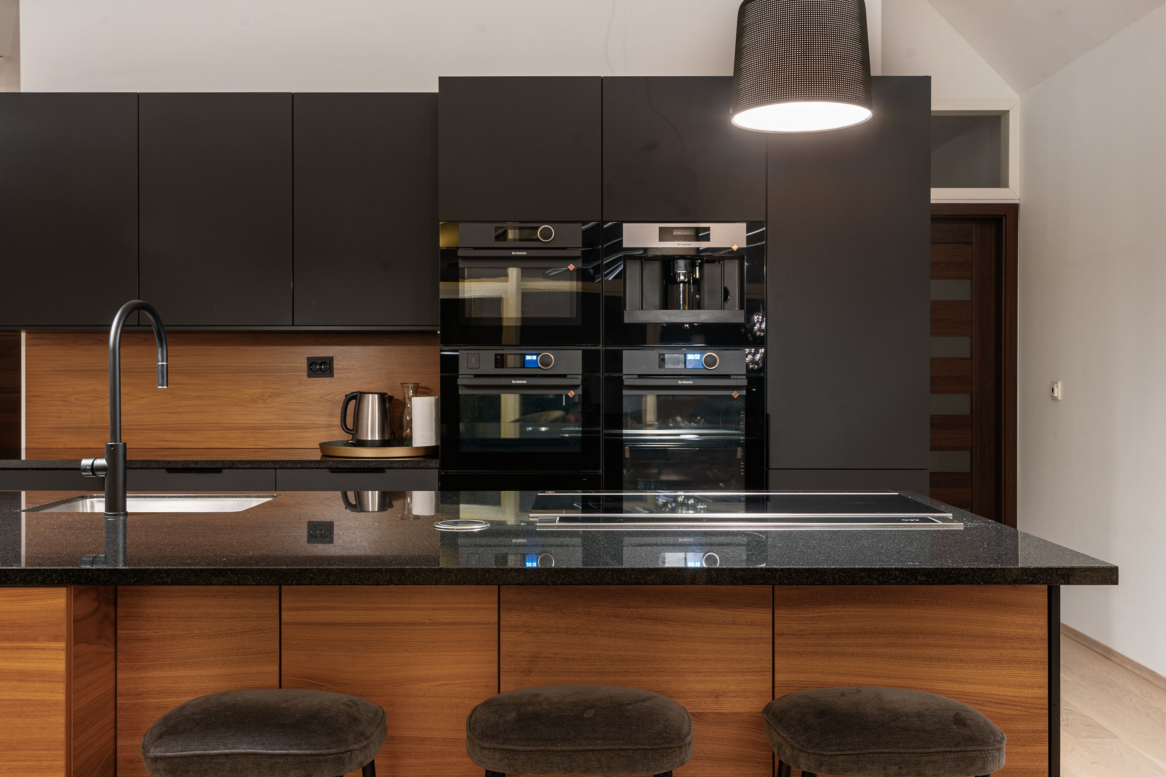 A modern kitchen with dark cabinetry, stainless steel appliances, a black countertop, and a pendant light.