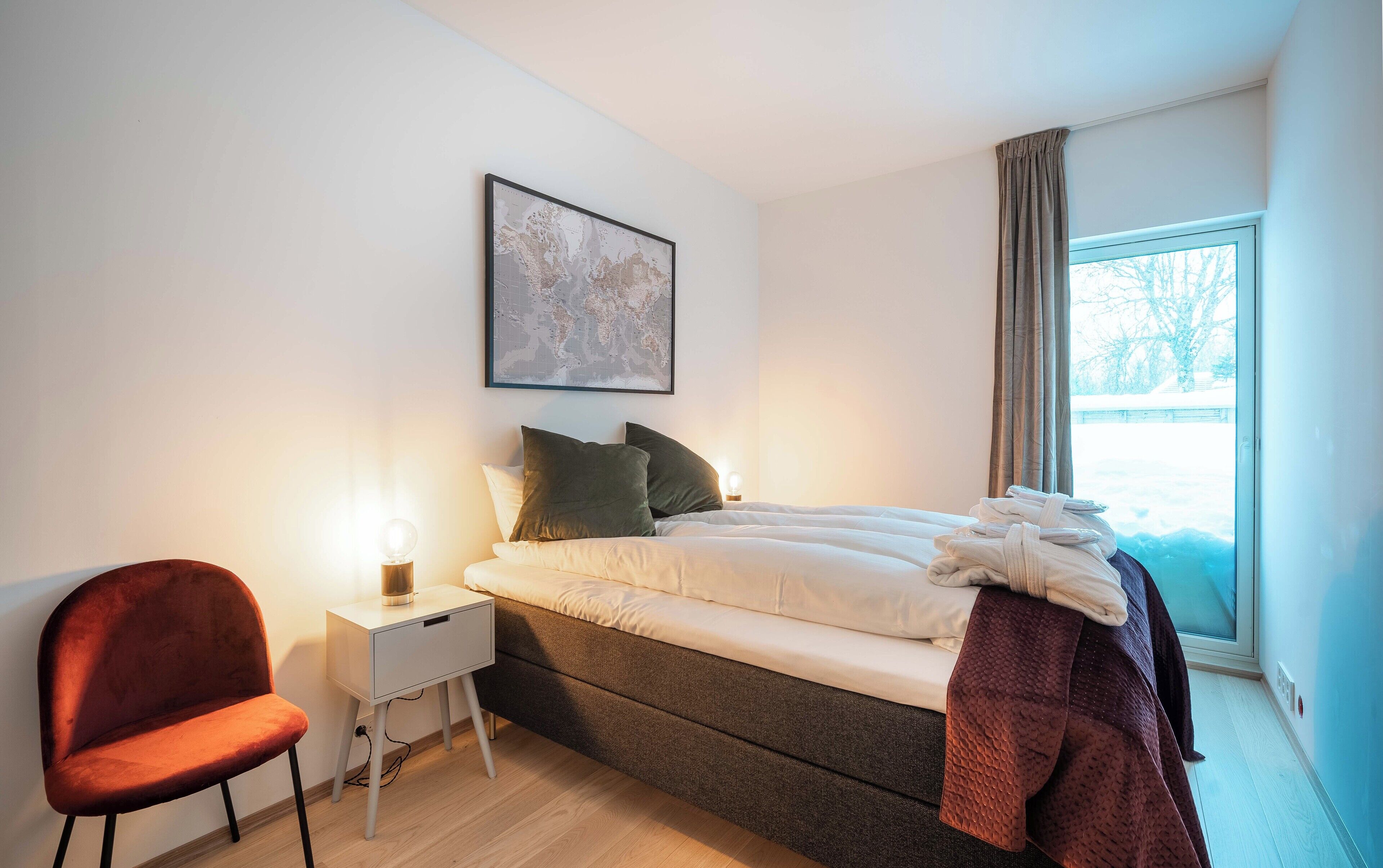 A bedroom with a bed, a nightstand, a chair, a map on the wall, and a window with curtains.