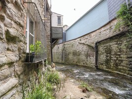 Un estrecho callejón con un muro de piedra, un pequeño arroyo y un edificio con una ventana y barandilla de metal.