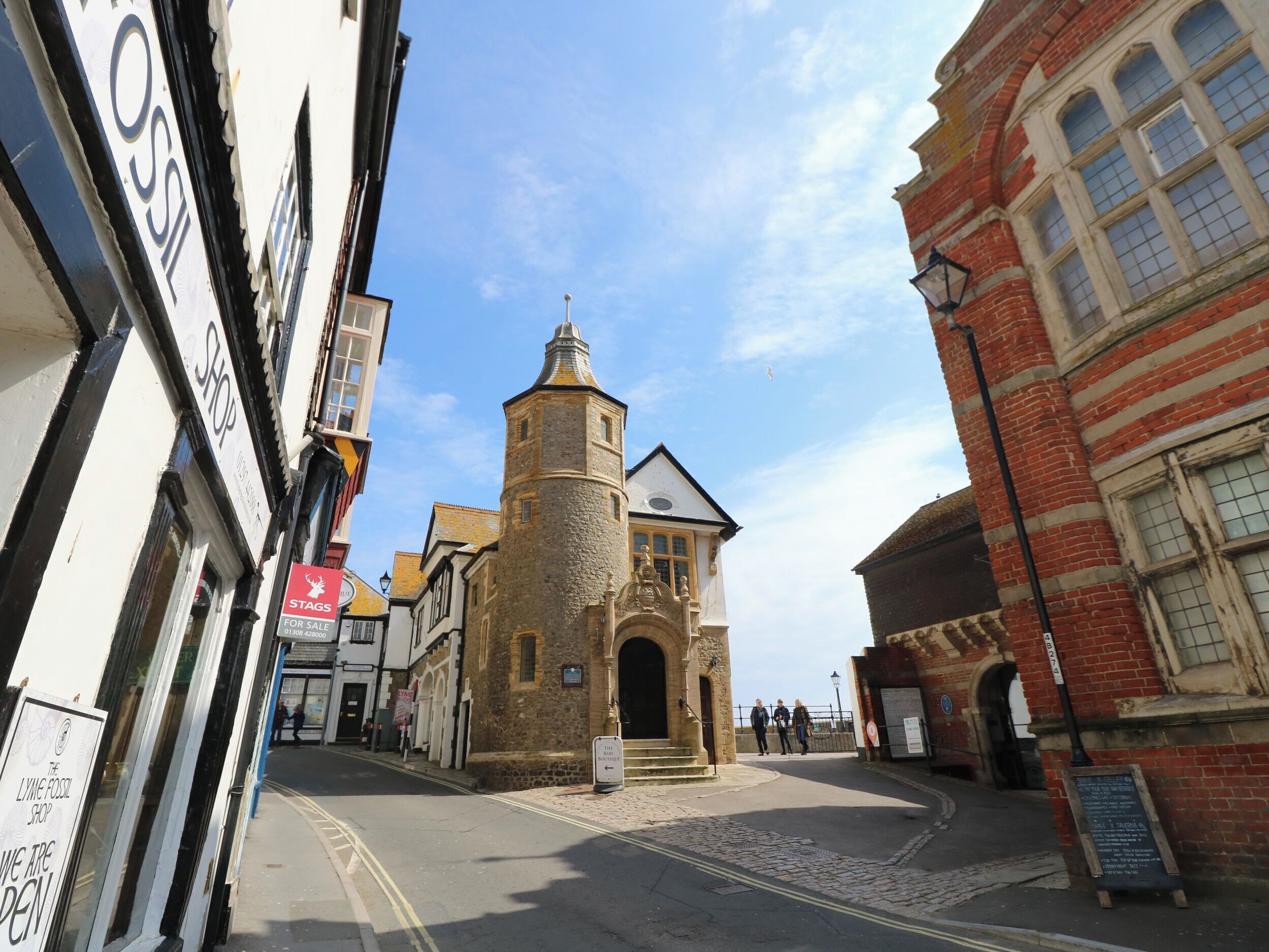 A cobblestone street with historic buildings, including a tower with a pointed roof and a brick building with arched windows.