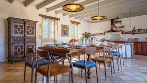 A dining area with a wooden table set for six, surrounded by upholstered chairs. A large wooden cabinet is to the left, and a kitchen area is visible in the background.