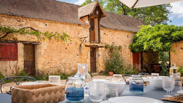 An outdoor dining area with a stone building, a wooden table set with plates, glasses, and a pitcher.