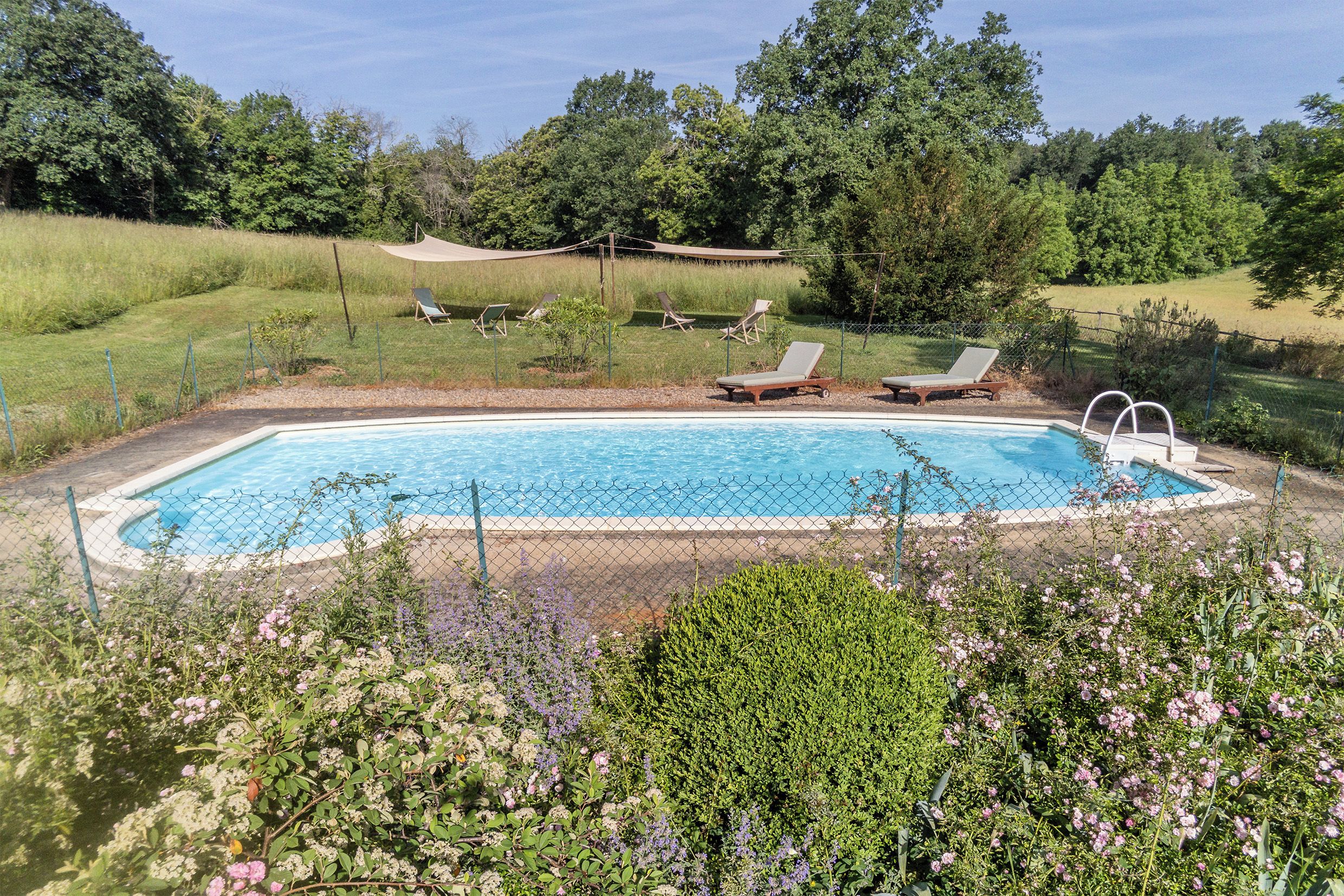 Ein rechteckiger Swimmingpool mit einem Zaun aus Maschendraht, umgeben von einer Rasenfläche mit mehreren Liegestühlen.