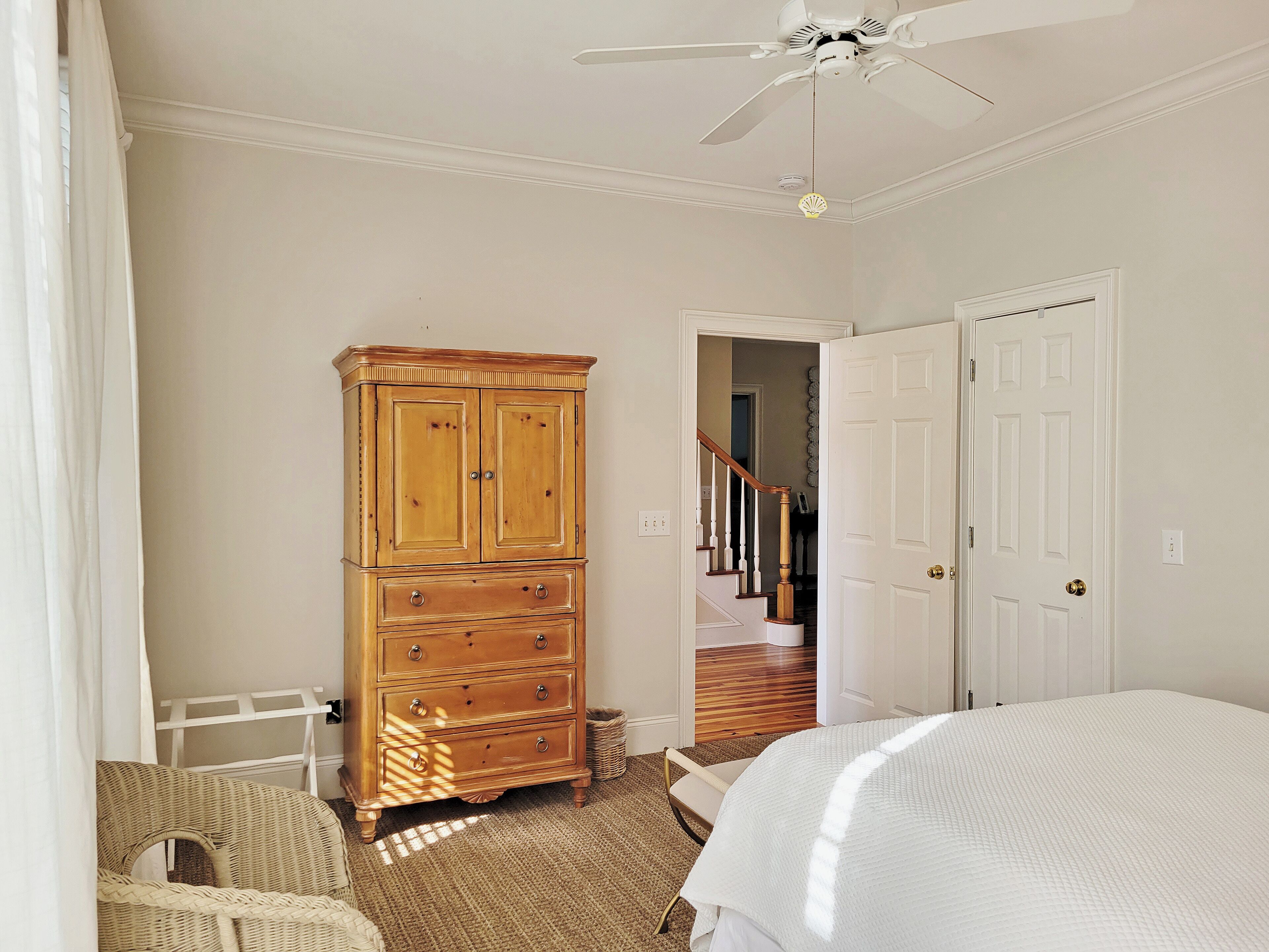 A bedroom with a wooden dresser, a bed, a ceiling fan, and a staircase leading to another room.
