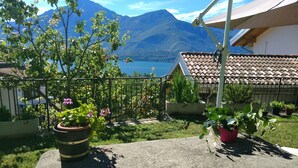 A balcony with potted plants, a view of a mountain and lake, and a tiled roof. - APARTMENT 1 BEDROOM WITH A FANTASTIC VIEW OF LAKE COMO (Gravedona Ed Uniti)