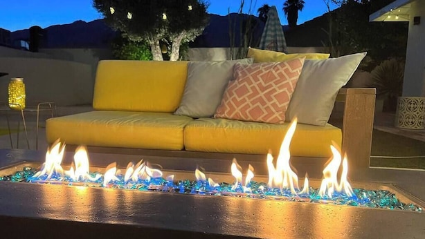 A fire feature with blue glass pebbles, a yellow sofa with patterned cushions, and a decorative lantern in a patio setting. - Amarillo House - Luxury Home With Pool Spa (Palm Springs)