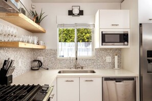 Una cocina moderna con armarios blancos, lavavajillas de acero inoxidable, microondas y ventana con vistas a zonas verdes.