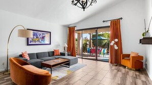 A modern living room with a sofa, armchair, and coffee table, a painting on the wall, and a view through a glass door to an outdoor area. - Sunset House (Palm Springs)