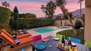 A poolside area with lounge chairs, a table set for dining, and a view of the pool and mountains. - Sunset House (Palm Springs)