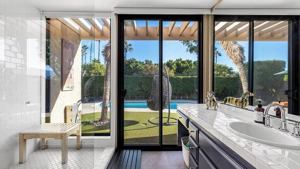 A modern bathroom with a large window, a wooden bench, a sink, and a shower area. - Sunset House (Palm Springs)