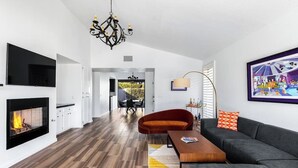A modern living room with a fireplace, a sofa, a coffee table, and a dining area visible in the background. - Sunset House (Palm Springs)