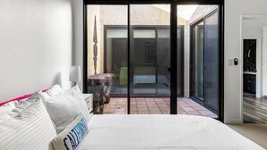 A bedroom with a bed, a nightstand, a lamp, and a sliding glass door leading to a patio. - Sunset House (Palm Springs)