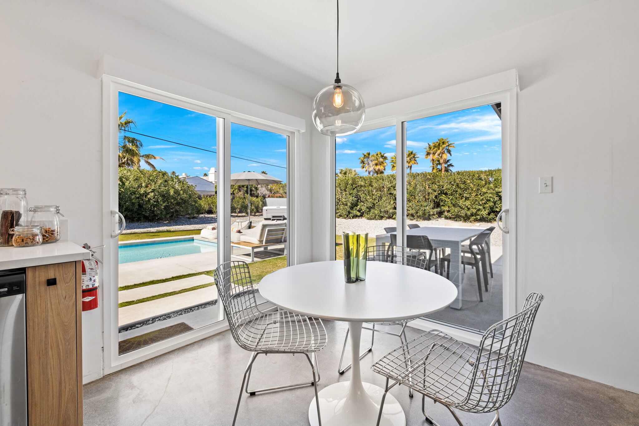 Un espace repas moderne avec une table blanche et des chaises en fil de métal, donnant sur une piscine et une végétation luxuriante.