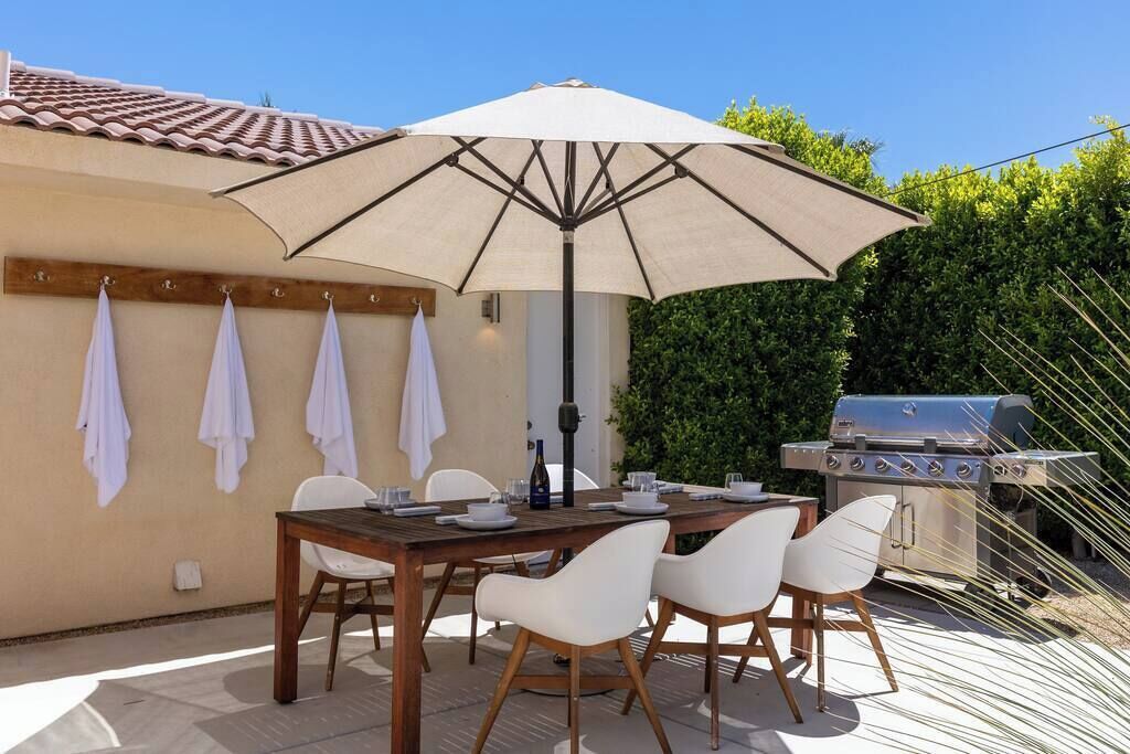 An outdoor patio with a wooden table, white chairs, a large umbrella, and a barbecue grill.