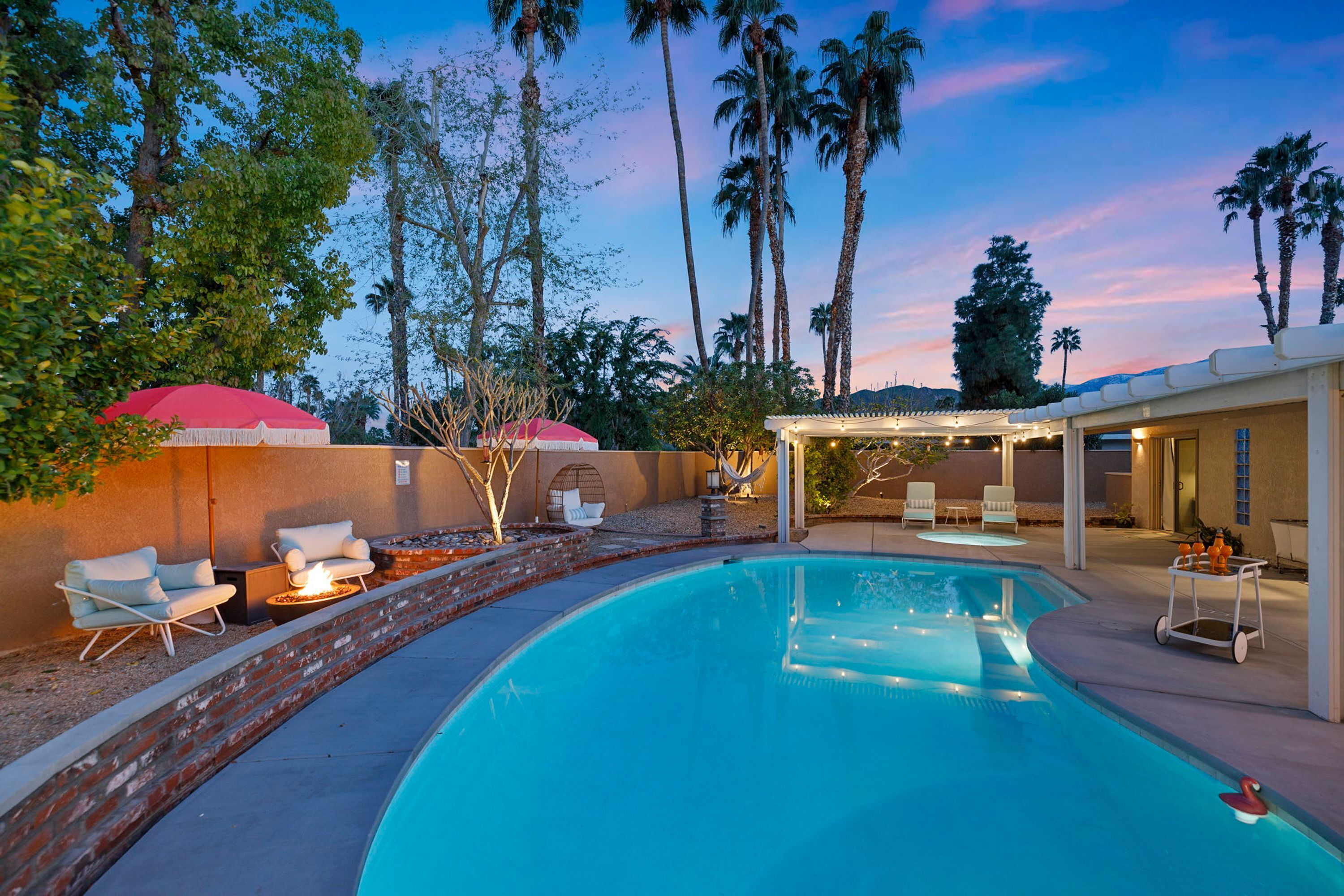 A swimming pool with lounge chairs and a covered seating area, surrounded by palm trees and a sunset sky.