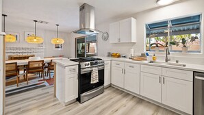 A modern kitchen with white cabinets, a built-in oven, and a dining area with a table and chairs. - The Fig Ranch (Palm Springs)