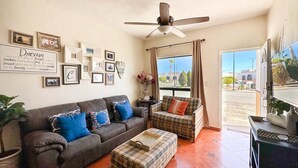 Un salon comprenant un canapé, un pouf, un ventilateur de plafond et une grande fenêtre avec des rideaux.
