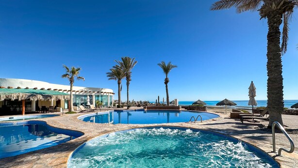 A resort pool area with multiple pools, including a hot tub, surrounded by palm trees and lounge chairs. - Breathtaking Luxury Oceanfront Condo (Puerto Peñasco)