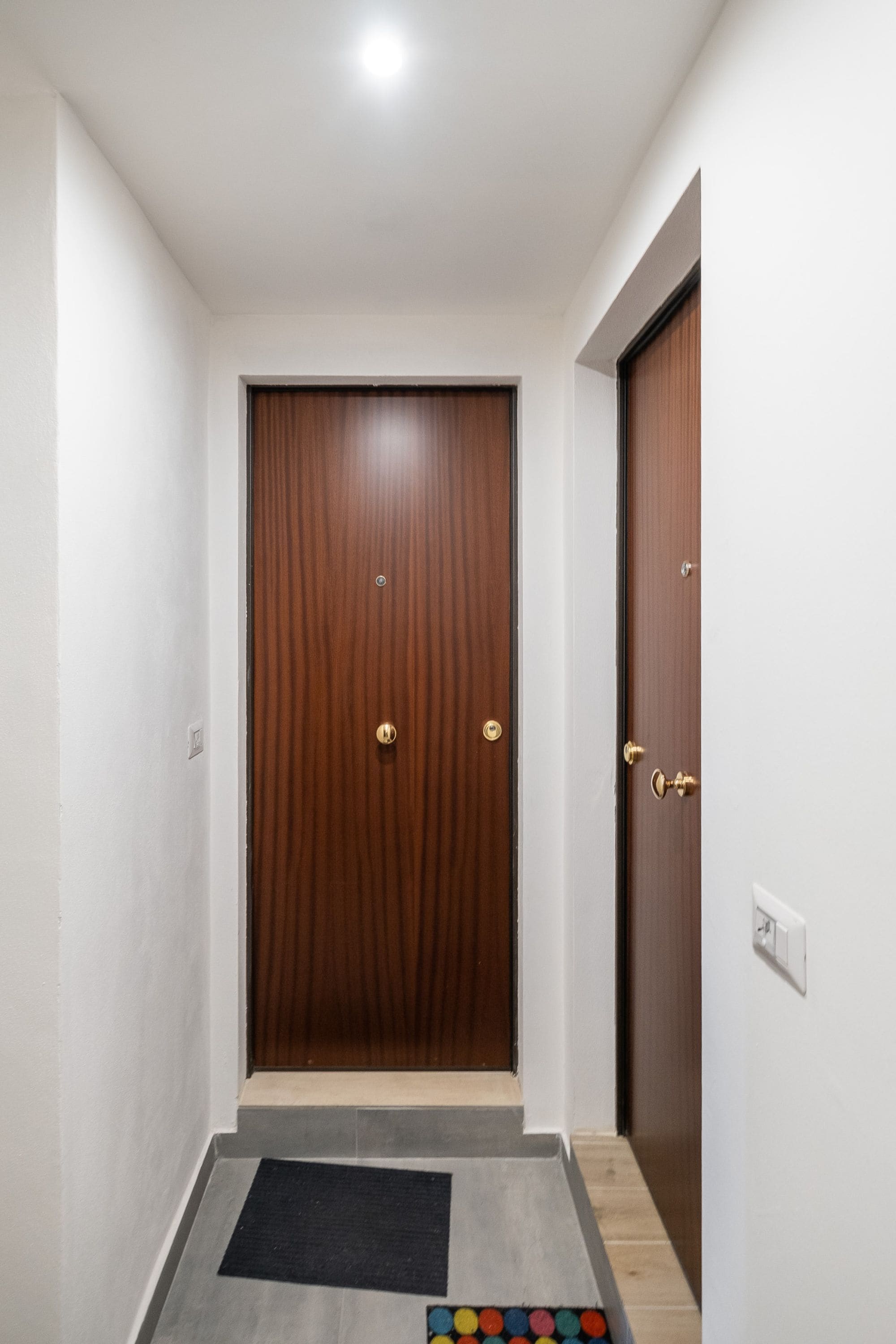 Un couloir avec une porte en bois, un petit paillasson et un tapis d’entrée orné de cercles colorés.