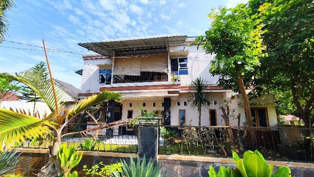 Une maison à deux étages dotée d’un balcon, entourée de plantes tropicales et surplombant un ciel dégagé.