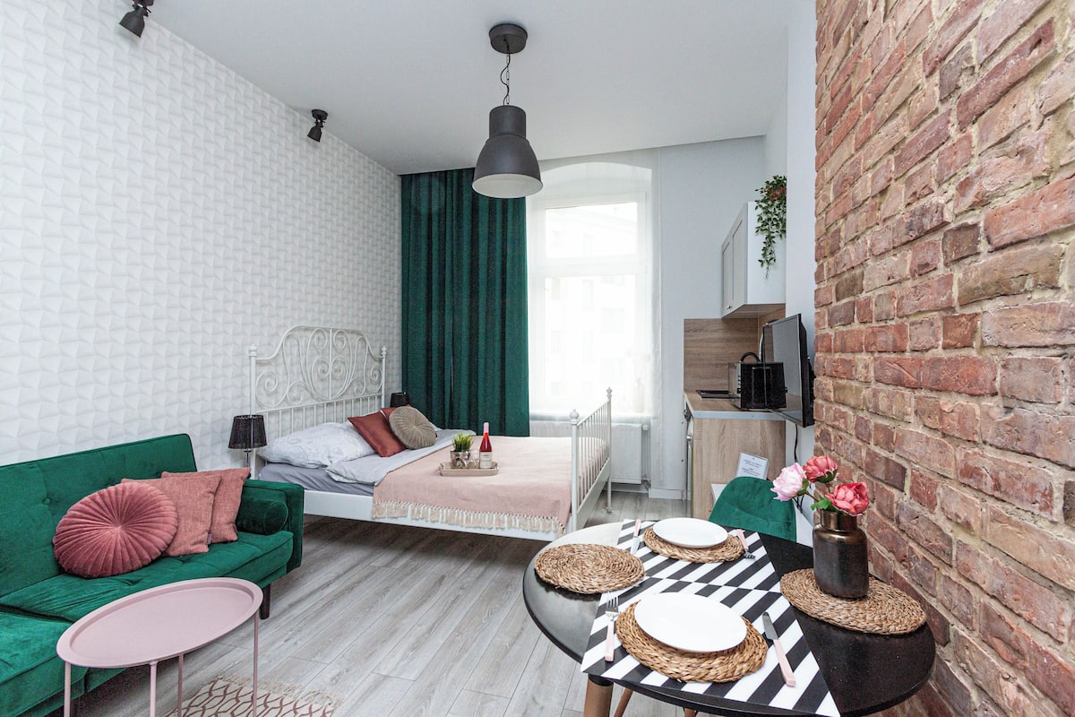 Un espace de vie moderne et compact, avec un canapé vert, une petite table ronde, un lit doté d’une tête de lit blanche et un mur en briques.
