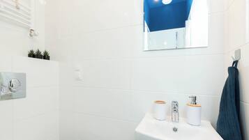Une salle de bain avec des carreaux blancs, un lavabo doté de deux distributeurs, un miroir et un porte-serviettes.