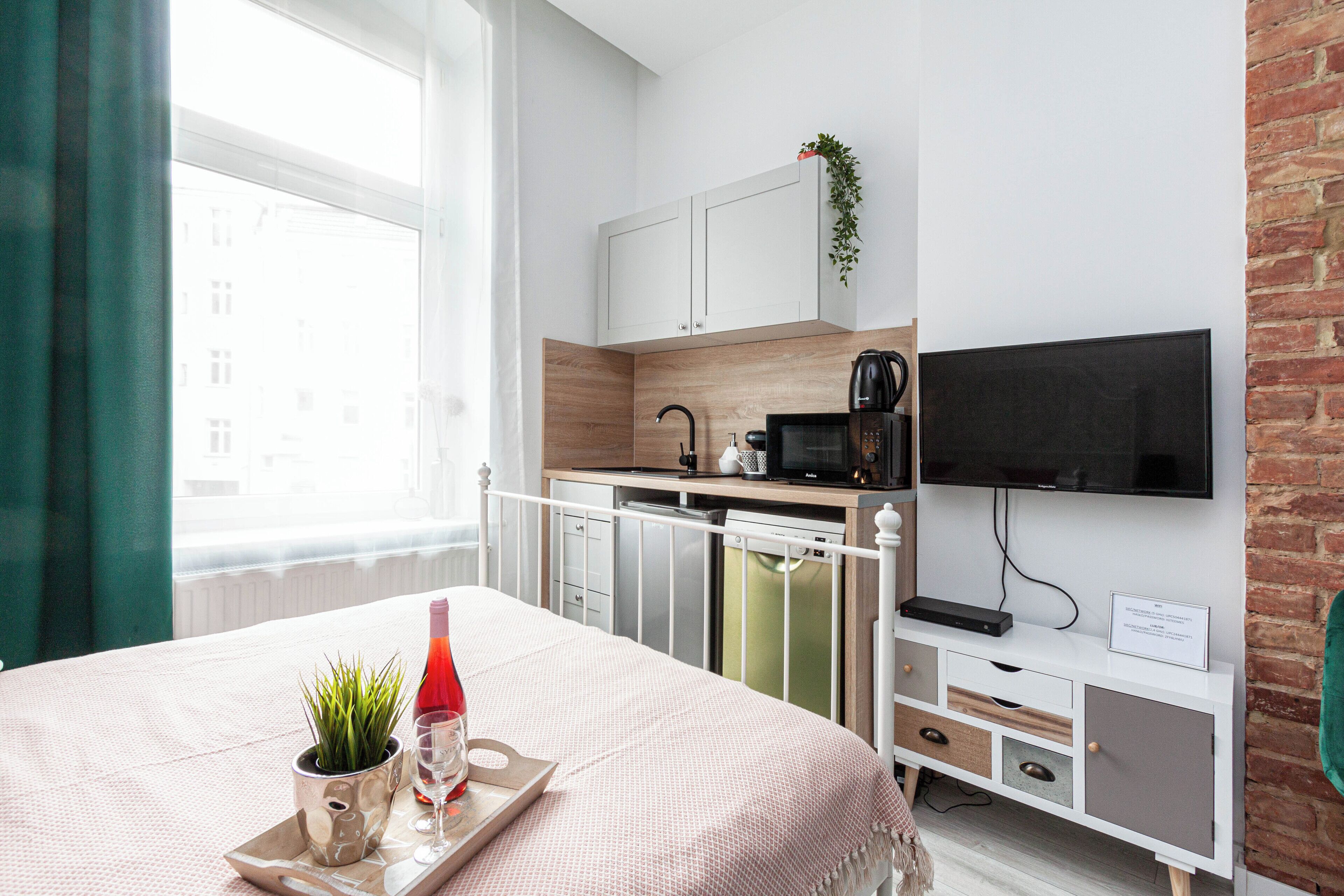 A modern hotel room with a bed, a small kitchenette, a TV, and a window with curtains.