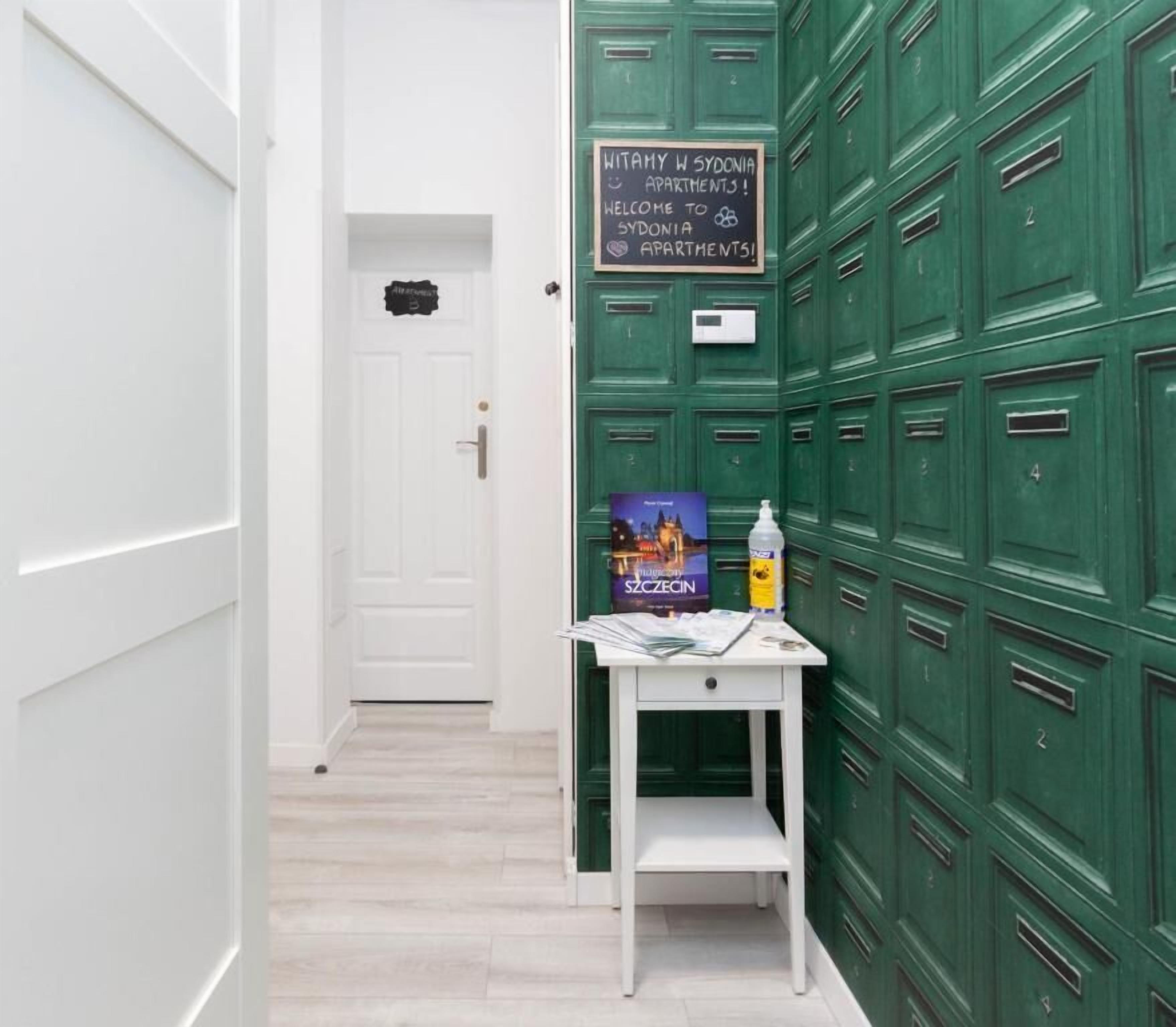 Un pasillo con taquillas verdes, una puerta blanca y una pequeña mesa con un cartel de bienvenida y un libro.