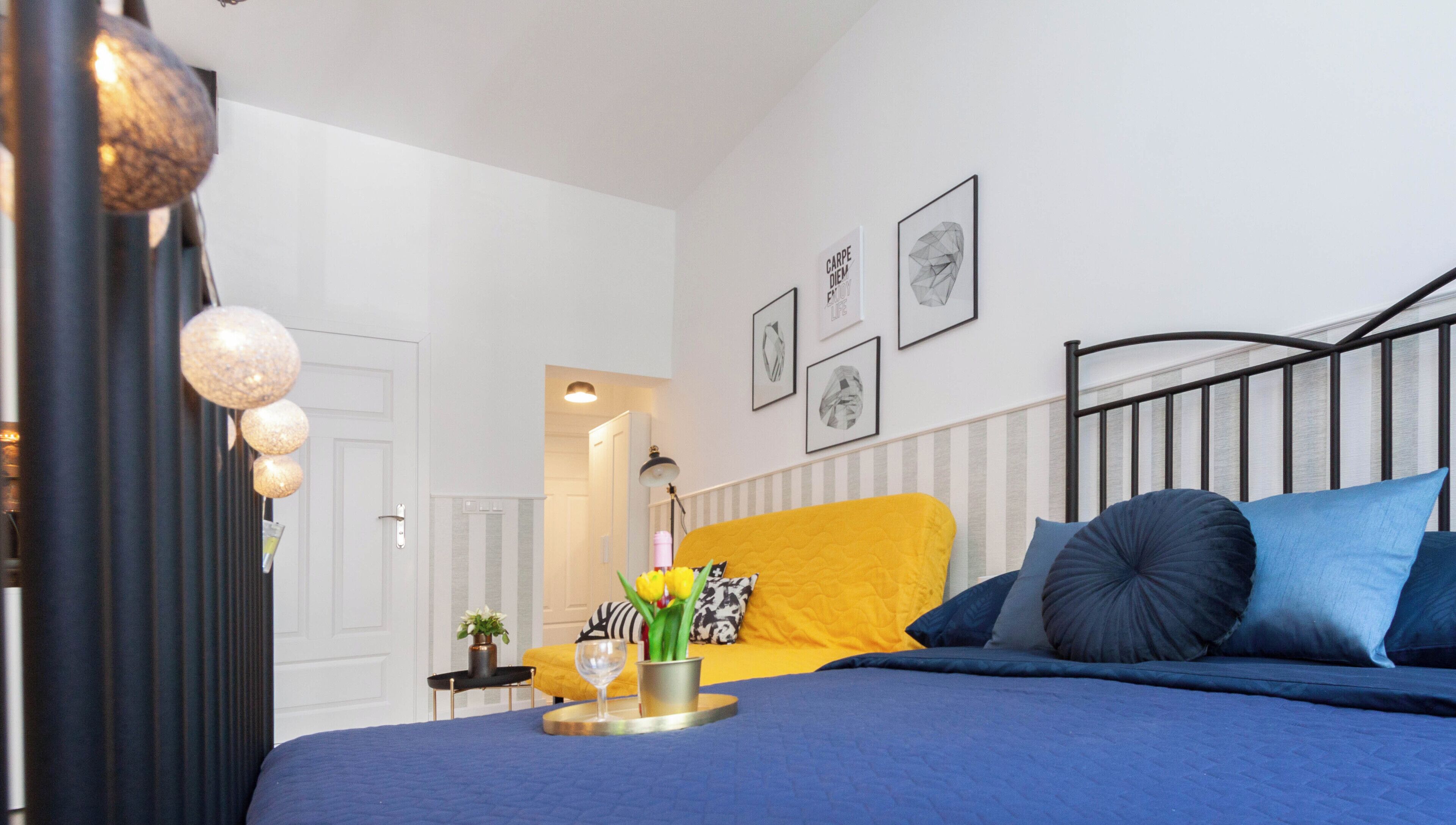 A bedroom with a bed, a bedside table, a lamp, and a wall with framed pictures.