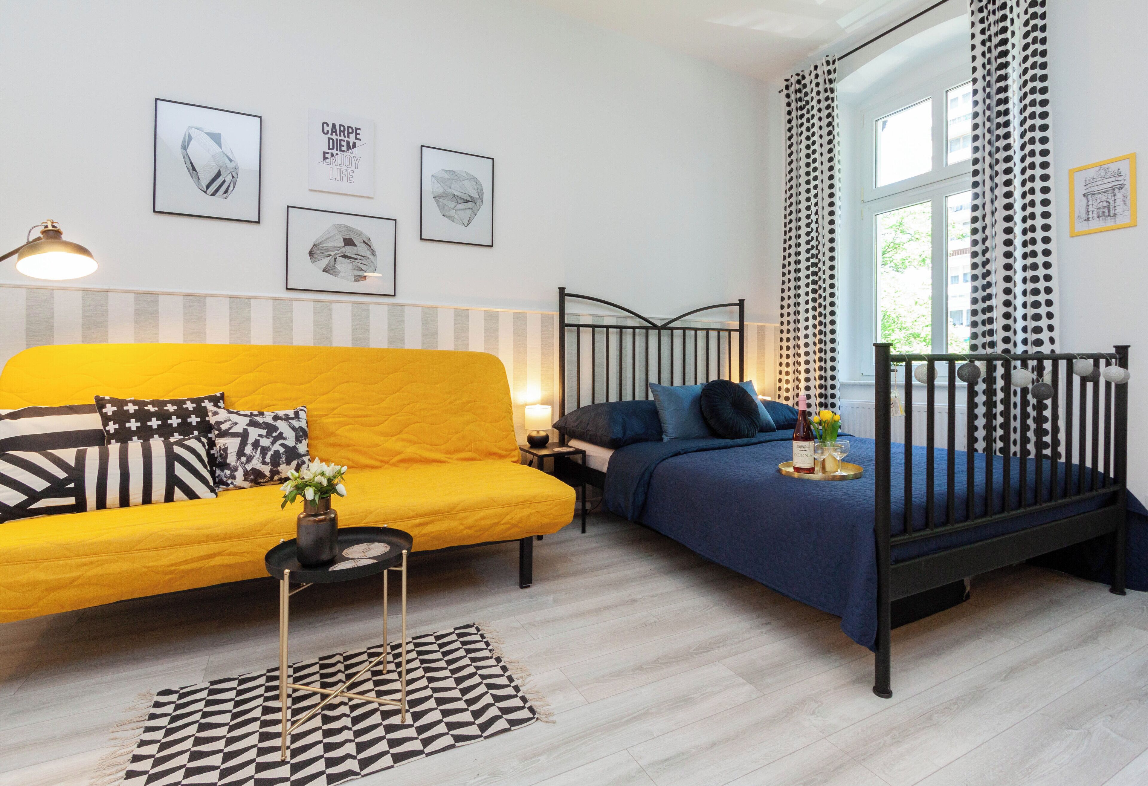 Una habitación con un sofá amarillo, una cama azul con estructura de metal negro, un tapete con estampado blanco y negro, y una mesita de noche con una lámpara.