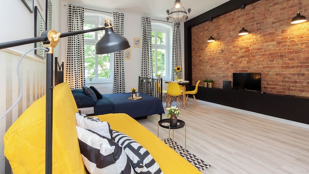 Una sala de estar moderna con un sofá amarillo, almohadas estampadas en blanco y negro, una lámpara de pie negra y una pared de ladrillo.