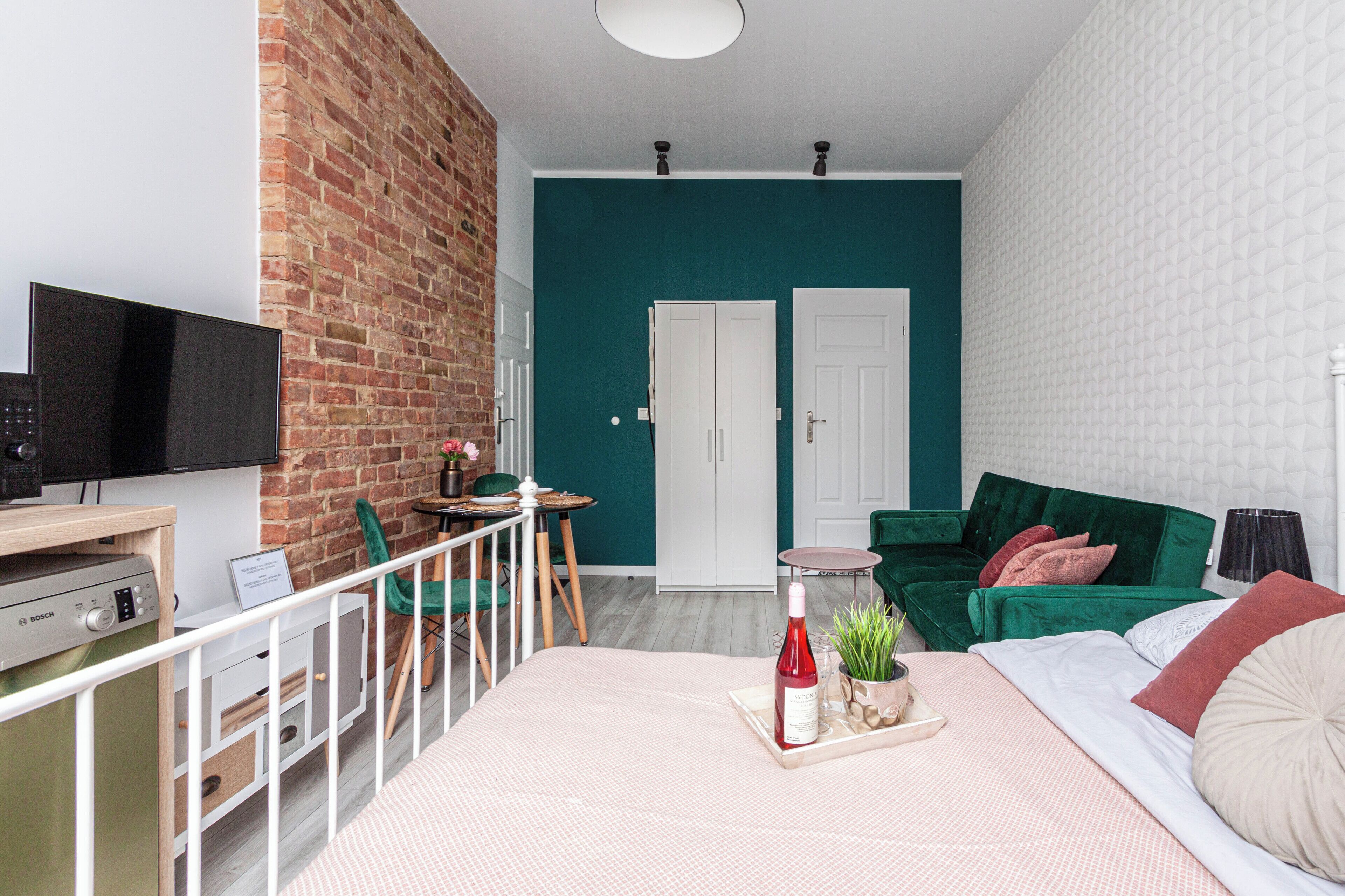Una habitación moderna con pared de ladrillo, sofá verde, cama con cabecera blanca, televisor de pantalla plana y una pequeña mesa con sillas.