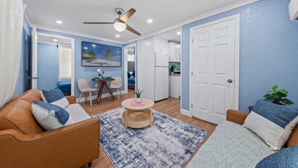 A modern living room with a brown leather sofa, a round coffee table, and a blue patterned rug. There is a dining area with a small table and chairs, and a kitchen visible in the background. - Cozy & Welcoming Home in Antique Row WPB (West Palm Beach)