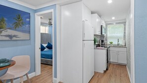 A compact hotel room with a kitchenette, a bed with blue bedding, a dining area with a table and chairs, and a painting of a beach scene on the wall. - Cozy & Welcoming Home in Antique Row WPB (West Palm Beach)