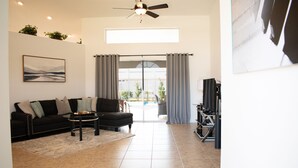Un salon avec un canapé noir, un fauteuil noir, une table basse et un meuble pour la télévision. On y trouve également un ventilateur de plafond, une fenêtre avec des rideaux et une porte donnant sur un espace extérieur.