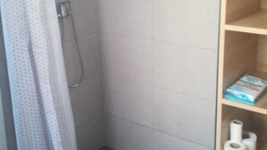 Ein Duschbereich mit gefliestem Boden, einer Duschbrause und einem Duschvorhang. Eine Holzablage mit einer Schachtel und zwei Toilettenpapierrollen.