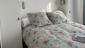 A bedroom with a bed, a nightstand, a shelf with books and a plant, and a window with curtains.